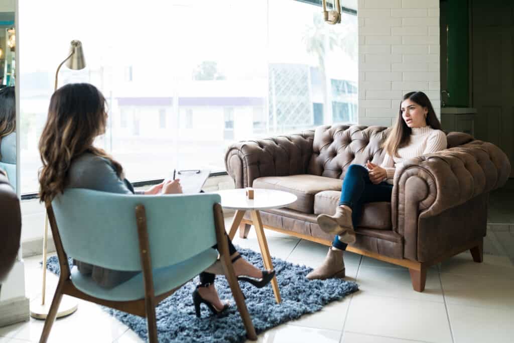 woman sitting on a couch talking to her therapist who is taking notes on a clipboard
