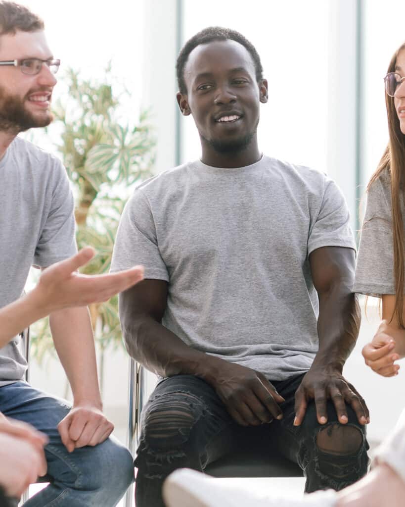 A man sitting on a chair in a therapy circle with another man next to him and a woman on the other
