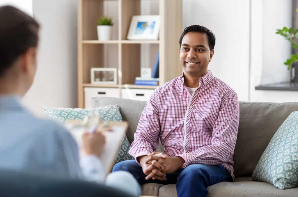 Smiling man sitting on a couch looking at his therapist who is sitting holding a notebook and pen writing things down